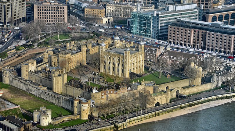 The Tower of London surrounded by ancient stone walls and the River Thames in the background on a sunny day.